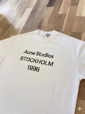 Vit Acne Studios t-shirt Stockholm - Vit t-shirt från Acne Studios med svart tryck framtill där det står 'Acne Studios STOCKHOLM 1996'. Klassisk rund halsringning och rak passform. Perfekt för dig som gillar minimalistisk och stilren design.