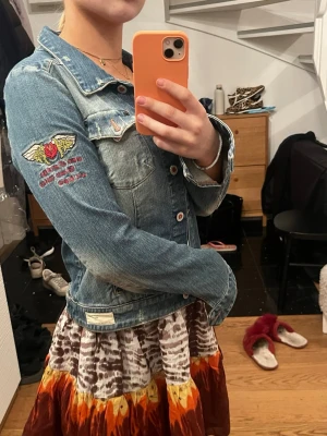 Blå jeansjacka med brodyr från odd molly - Snygg blå jeansjacka med cool brodyr på ärmen som föreställer en röd ros med vingar och text. Klassisk modell med bröstfickor och silverfärgade knappar. Perfekt att slänga över en klänning eller t-shirt för en avslappnad look. 