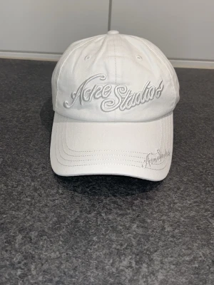 Vit keps från Acne Studios - Säljer en stilren vit keps från Acne Studios med broderad logga framtill och på skärmen. Klassisk böjd skärm och justerbar baksida. Perfekt accessoar för en clean och trendig look. Materialet är bomull.