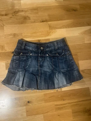 Mörkblå lågmidjad kort jeanskjol - Kort jeanskjol i mörkblå denim med slitna kanter och veckad design. Kjolen har fram- och bakfickor, bälteshällor och knäppning fram. Perfekt för en avslappnad och trendig look. Den är lågmidjad och kort. Säljer för att jag växt ut. 