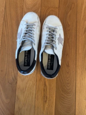 Golden Goose vita sneakers med stjärna - Golden Goose sneakers i vitt läder med grå mockastjärna på sidan och grå skosnören. Svart detalj vid hälen och texten 'SUPER-STAR' på innersulan. Klassisk låg modell med slitna detaljer för en cool, avslappnad look.
