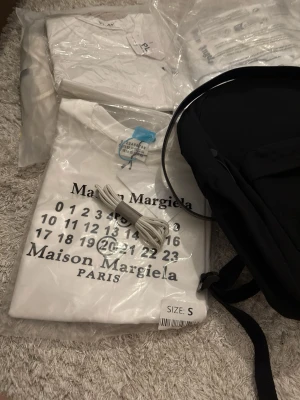 Maison Margiela vit t-shirt - Vit t-shirt från Maison Margiela med det ikoniska siffertrycket och loggan på bröstet. Klassisk passform och rund halsringning. Tillverkad i mjuk bomull som känns skön mot huden. Perfekt för dig som gillar stilrena och trendiga plagg.