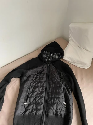 Svart cardigan  - Snygg svart hoodie med quiltad front och dragkedja. Utrustad med huva som har rutigt foder och dragsko. Ärmar och rygg i mjukt tyg, medan framsidan är vadderad för extra värme. Två fickor med dragkedja framtill. Perfekt för en cool och avslappnad stil.
