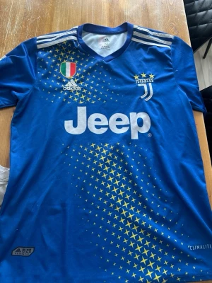 Blå Juventus matchtröja Adidas - Juventus matchtröja i blått med gula stjärnor, vita detaljer och klubbmärke. Adidas-logga och Jeep som sponsor på bröstet. Tröjan har vita ränder på axlarna och är tillverkad i lätt, ventilerande Climalite-material. Perfekt för fotbollsträning eller support.
