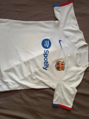 Vit FC Barcelona matchtröja Nike - Säljer en vit FC Barcelona matchtröja från Nike med Spotify-logga på bröstet. Tröjan har klubbmärket broderat, blå Nike-logga och detaljer i blått och rött på ärmsluten. Perfekt för dig som älskar fotboll och vill stötta Barca.