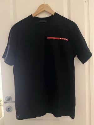 Svart t-shirt från Prada - Helt ny äkta svart Prada T-shirt med prislappen kvar. Storlek S. Normal passform. Mått hänvisas på sista bilden. Slutsåld på deras hemsida. 