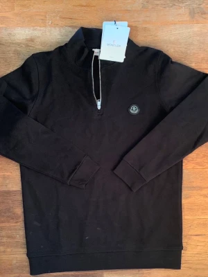 Svart halfzip hoodie från Moncler - Svart hoodie från Moncler med halv dragkedja och Moncler-logga på bröstet. Tillverkad i mjukt bomullsmaterial och har ribbade muddar vid ärmslut och nederkant. Perfekt för en clean och stilren look.
