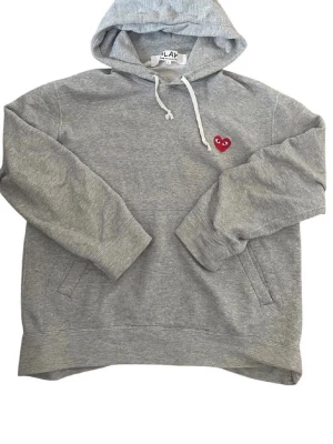 Grå hoodie Comme des Garçons Play - Grå hoodie från Comme des Garçons Play med huva och vita dragsnören. Klassisk röd hjärtlogga med ögon på bröstet. Tillverkad i mjuk bomullsblandning och har fickor på sidorna. Perfekt för en chill och stilren look.