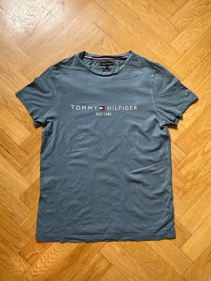 Blå/grå Tommy Hilfiger t-shirt - Snygg blå t-shirt från Tommy Hilfiger med tryckt logga och text 'EST. 1985' på bröstet. Klassisk passform med rund hals och korta ärmar. Tillverkad i mjuk bomull, perfekt för en avslappnad stil.