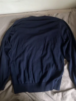 Mörkblå tröja från Lacoste - Mörkblå långärmad tröja från Lacoste med klassisk v neck och den ikoniska krokodilloggan broderad på bröstet. Tröjan har ribbade muddar vid ärmslut och nederkant. Perfekt för en stilren och enkel look. Pris kan snackas om.