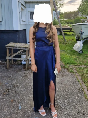 Marinblå långklänning med slits - Säljer en fin marinblå långklänning i ett glansigt material med hög slits framtill och knyt i midjan. Klänningen är ärmlös och har en rund halsringning, använd 2 gånger.