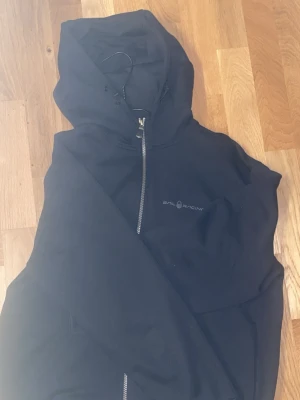 Svart hoodie från Sail Racing - Svart hoodie med dragkedja från Sail Racing. Klassisk modell med huva och diskret logga på bröstet. Perfekt för en avslappnad stil. Mjukt material och sköna muddar vid ärmslut och nederkant. Helt ny storlek M passsar L med 