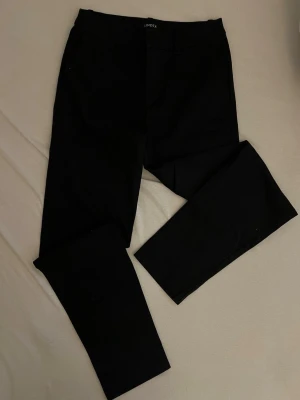 Svarta kostymbyxor från Lindex - Stilrena svarta kostymbyxor från Lindex med klassisk rak passform och hällor för skärp. Perfekta för en dressad look. Byxorna har diskreta fickor och är tillverkade i ett mjukt tyg som ger en snygg siluett.