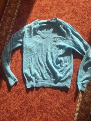 Ljusblå stickad tröja från Lyle & Scott - Ljusblå stickad tröja med V-ringning från Lyle & Scott. Tröjan har lång ärm och klassisk gul logotyp med fågel på bröstet. Perfekt för lager-på-lager och enkel att matcha med jeans eller kjol.