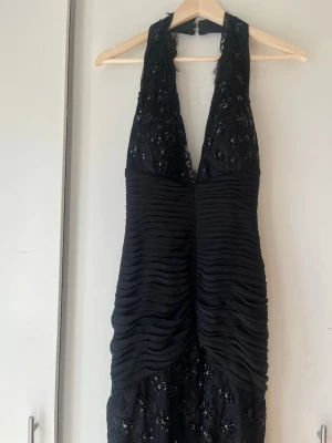 Svart långklänning med spets och pärlor - Elegant svart långklänning med djup v-ringning, dekorerad med spets och svarta pärlor upptill. Klänningen har ett plisserat midjeparti och är ärmlös med halterneck. Perfekt för en glamorös kväll.