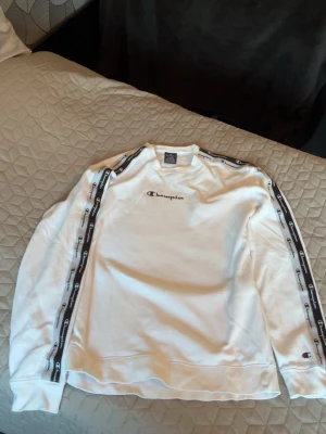 Vit Champion sweatshirt med logoband - Vit långärmad sweatshirt från Champion med svarta logoband längs ärmarna och broderad logga på bröstet. Klassisk crewneck-modell i mjukt material, perfekt för en avslappnad streetstyle-look.Storlek M