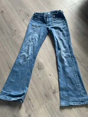 Blå utsvängda jeans från Gina Tricot - Snygga ljusblå jeans från Gina Tricot med utsvängda ben och markerade sömmar framtill. Jeansen har två fickor fram och dubbla silverknappar i midjan. Perfekta för dig som gillar retrostil och vill sticka ut lite extra.