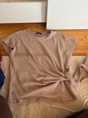 Beige stickad topp med knyt från Zara - Säljer en beige stickad topp från Zara med rund hals och korta ärmar. Toppen har en snygg knyt-detalj i sidan som ger en trendig look. Perfekt att styla med jeans eller kjol för en chill vibe.