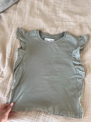 Grön ärmlös topp med volang - Snygg ljusgrön ärmlös topp från H&M med volangdetaljer längs axlarna. Perfekt för en trendig och avslappnad look. Mjuk bomullskvalitet och rund halsringning.