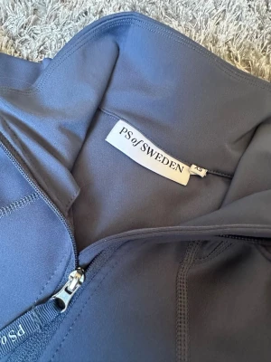 Blå/Grå polotopp från PS of Sweden (Nyskick) - Blå/Grå långärmad polotopp i en äldre modell från PS of Sweden med halv dragkedja framtill. Figurnära passform och tillverkad i stretchigt syntetmaterial, perfekt för träning och ridning. Använd fåtal gånger och ser ut som ny!❤️ (Det är bara att skriva om du har några frågor)