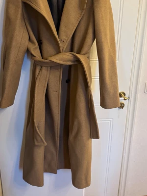 Mango kamelbrun lång rock med knytskärp - Säljer en stilren och klassisk kamelbrun rock med knytskärp i midjan och svarta knappar. Rocken har bred krage och är helfodrad med mörkt innerfoder. Perfekt för dig som vill ha en tidlös och elegant look under höst och vinter.