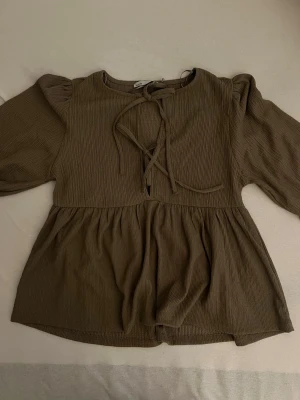 Brun blus med knytning fram - Snygg brun blus med kortärmade puffärmar och knytning framtill. Blusen har en lätt rynkad midja och är tillverkad i ett mjukt, ribbat tyg som ger en avslappnad men trendig look. Perfekt att styla med jeans eller kjol.