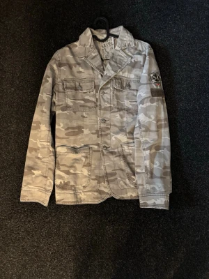  Sublevel, S field jacket  - Säljer en grå och beige camouflagejacka från Sublevel med coola badges på ärmen. Jackan har flera fickor framtill, knappar och klassisk krage. Ärmarna kan rullas upp och fästas med knapp. 