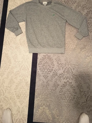 Grå sweatshirt från Lacoste - Klassisk grå sweatshirt från Lacoste med rund halsringning och ribbade muddar. Tröjan har det ikoniska gröna krokodilmärket broderat på bröstet. Perfekt för en avslappnad och stilren look.