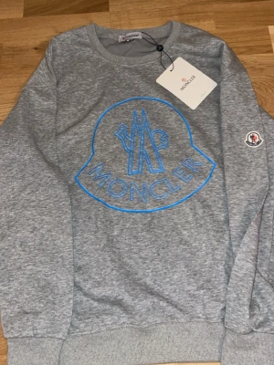 Moncler sweatshirt - Snygg grå sweatshirt från Moncler med stor blå logga framtill och Moncler-märke på ärmen. Tröjan har rund halsringning och ribbade muddar vid ärmslut och nederkant. Perfekt för en trendig och avslappnad stil.