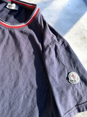 Mörkblå Moncler t-shirt med logga - Snygg mörkblå t-shirt från Moncler med klassisk logga på ärmen. T-shirten har rund halsringning med röd och vit randig kant. Tillverkad i mjuk bomull och har en normal passform. Perfekt för en clean och stilren look. 