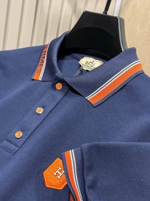 Pikétröja  - Snygg marinblå pikétröja med orange och vita detaljer på krage och ärmslut. Tröjan har knappar framtill och ett orange broderat H-märke på bröstet. Perfekt för en stilren och sportig look.