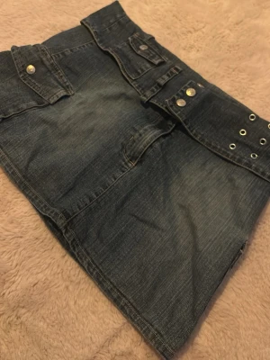 Mörkblå mini jeanskjol med fickor - Cool mörkblå jeanskjol i klassisk denim med två stora fickor framtill och snygga silverfärgade knappar och nitar. Kjolen har en rak passform och detaljer med öljetter i midjan. 