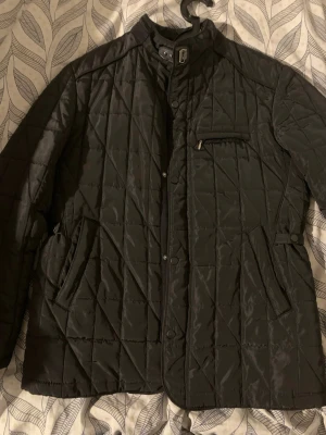 Svart grisch jacka med ståkrage - Snygg svart quiltad jacka med ståkrage och tryckknappar. Jackan har dragkedja och knäppning framtill, två snedställda fickor samt en bröstficka med dragkedja. Justerbara spännen i midjan och diskret rutmönster i tyget.