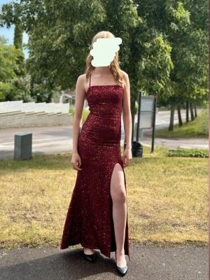 Glittrig vinröd långklänning - Snygg vinröd långklänning med glitter, figurnära passform och hög slits framtill. Klänningen har smala axelband och öppen rygg med korsade band. Perfekt för dig som vill sticka ut på bal eller fest. Materialet är glittrigt och lite stretchigt för en bekväm känsla. Nypris 460