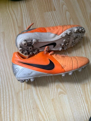 Nike orange fotbollsskor med dobbar - Säljer ett par orangea Nike fotbollsskor med svarta detaljer och silverfärgad häl. Skorna har klassisk snörning och gummidobbar för bra grepp på planen. Ovandelen är i syntetmaterial med textur för bättre bollkontroll. Perfekta för dig som vill sticka ut på planen.