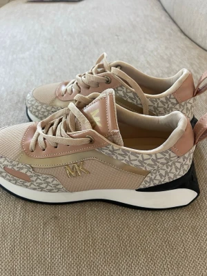 Beige sneakers från Michael Kors - Snygga sneakers i beige och ljusrosa från Michael Kors med guldfärgade MK-detaljer på sidan. Skorna har mönstrad textil, mesh och läderdetaljer samt vit och svart sula. Perfekta för dig som gillar stilrena och trendiga sneakers.