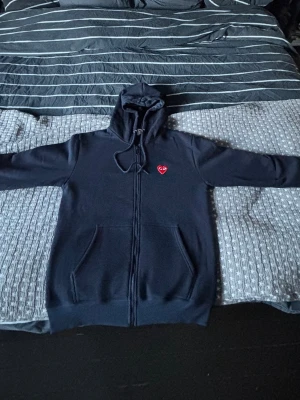 Mörkblå hoodie Cdg play - Svart hoodie med dragkedja från Cdg Pla, Klassisk design med huva, två fickor fram och det ikoniska röda hjärtat broderat på bröstet. Perfekt för en stilren och avslappnad look.