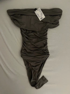 Grå off-shoulder body H&M  - Trendig grå body från H&M med off-shoulder design och rynkade detaljer längs sidorna. Tillverkad i ett stretchigt, mjukt material som sitter snyggt på kroppen. Praktisk knäppning i grenen för enkel användning. Perfekt för att skapa en stilren look. Och ser fantastisk ut i kostymbyxor 🙌