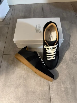  Maison Margiela Gats  - Snygga svarta sneakers från Maison Margiela med klassisk design, vita snören och kontrasterande gummisula. Ovandelen är i mocka och läder, och insidan har en beige ton. Perfekta för dig som gillar stilrena och tidlösa skor.