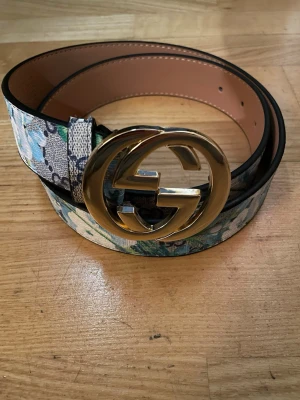 Blommigt skärp med guldspänne - Säljer ett snyggt skärp med blommigt mönster i blått, grönt och vitt. Skärpet har ett stort, rundat guldspänne med GG-logga. Perfekt för att lyfta din outfit med en exklusiv touch.