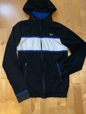 Svart vindjacka från Lacoste - Snygg svart vindjacka från Lacoste med vit och blå rand över bröstet. Jackan har huva, dragkedja framtill och två fickor med dragkedja. Klassisk Lacoste-logga på bröstet och blå detaljer vid ärmsluten. Perfekt för en sportig och stilren look.