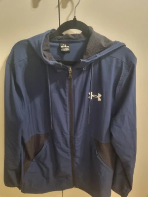 Blå träningsjacka från Under Armour - Snygg blå träningsjacka från Under Armour med dragkedja framtill och huva. Jackan har svarta mesh-detaljer på sidorna och innerfodret i huvan är också i mesh. 