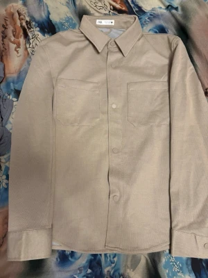 Beige overshirt från Zara - Stilren beige overshirt från Zara med klassisk krage, två bröstfickor och knäppning framtill. Overshirten har långa ärmar med manschett och är tillverkad i ett mjukt tyg som ger en avslappnad look.