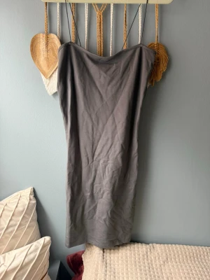Grå axelbandslös klänning - En enkel och stilren grå klänning med smala axelband och axelbandslös design. Klänningen har en rak och kroppsnära form som passar perfekt för en modern look. Materialet känns mjukt och bekvämt, idealiskt för att skapa en avslappnad men snygg outfit.