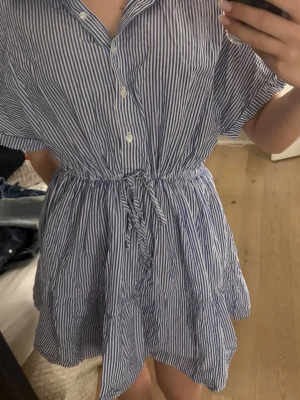Blåvit randig kortklänning med knyt - Säljer en blåvit kortklänning med tunna vertikala ränder, knappar framtill och knyt i midjan. Klänningen har korta ärmar, krage och avslappnad passform. Perfekt för varma dagar och enkel att styla med sneakers eller sandaler.