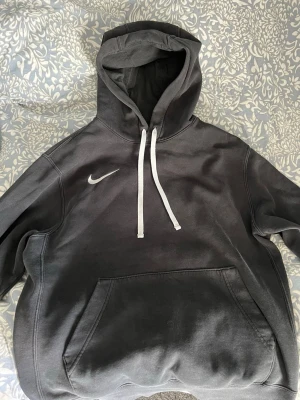 Svart Nike hoodie med huva - Svart hoodie från Nike med vit logga på bröstet och vita dragsnören i huvan. Klassisk känguruficka framtill och mjukt material som känns skönt mot huden. Perfekt för en avslappnad och sportig stil.