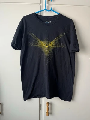 Svart Marvel t-shirt med gul tryck - Cool svart t-shirt från Marvel med ett unikt gult tryck av en karaktär på bröstet. Tillverkad i 100% bomull och har en klassisk passform med korta ärmar. Perfekt för dig som gillar superhjältar och vill sticka ut med en snygg grafisk design.