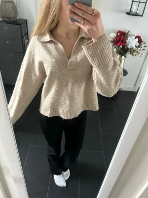 Beige stickad tröja med krage - Mysig beige stickad tröja med krage och v-ringning. Tröjan har långa ärmar och ribbstickade muddar. Som man kan se på bilden är den lite bopprig och sömmen har även gått upp på ena mudden (se bild 2)