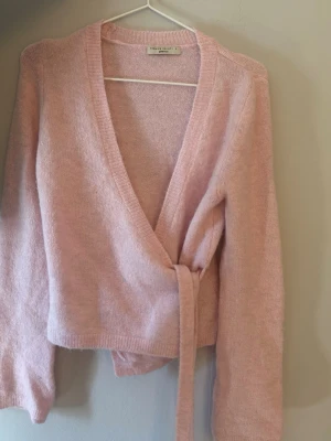 Ljusrosa omlottkofta från Gina - Mysig ljusrosa omlottkofta från Gina med knytband i midjan och långa ärmar. Koftan är stickad i ett mjukt material och har en djup V-ringning. Perfekt för lager-på-lager och en söt touch till din outfit.