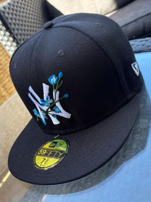 Svart New Era NY keps med blommor - Säljer en svart New Era 59FIFTY keps med broderad vit NY-logga och blå blommor på framsidan. Kepsen har rak skärm och klassisk passform. Materialet är bomull och loggan finns även broderad baktill. Perfekt för dig som vill sticka ut med snygga detaljer.